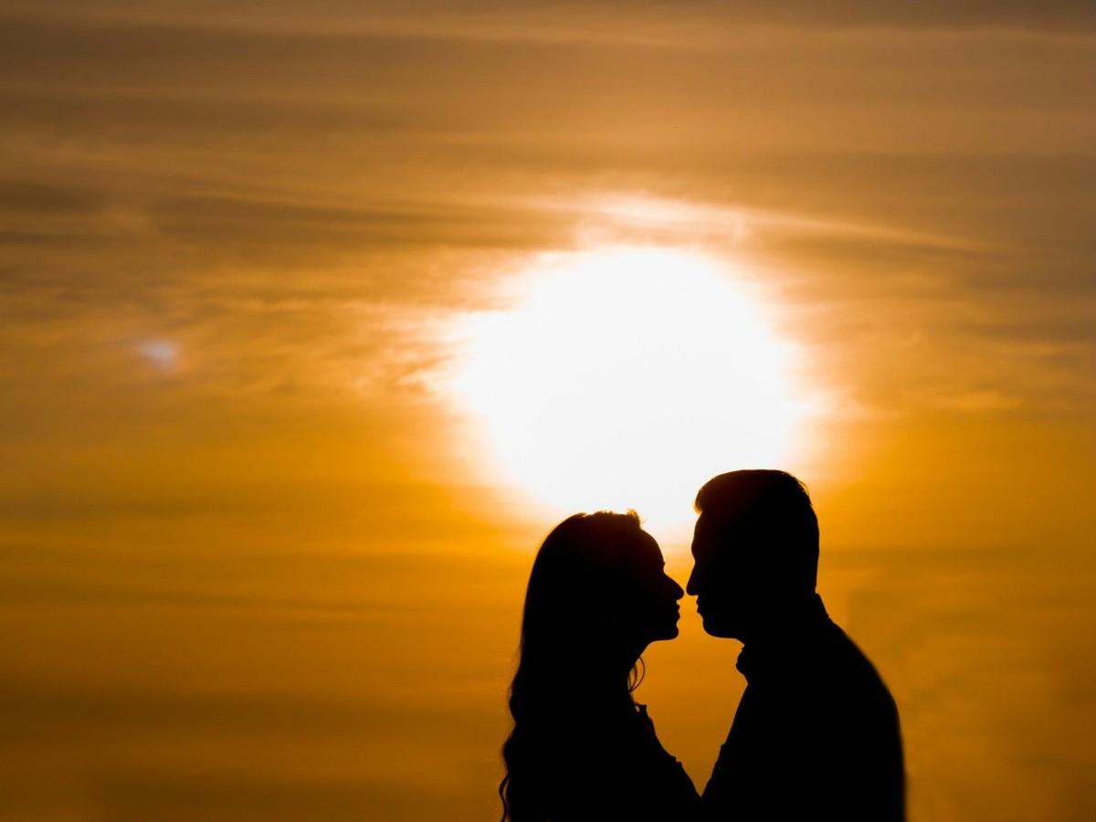 Silhueta de um homem e uma mulher frente a frente, muito próximos, contra um pôr do sol alaranjado intenso, clima romântico e acolhedor